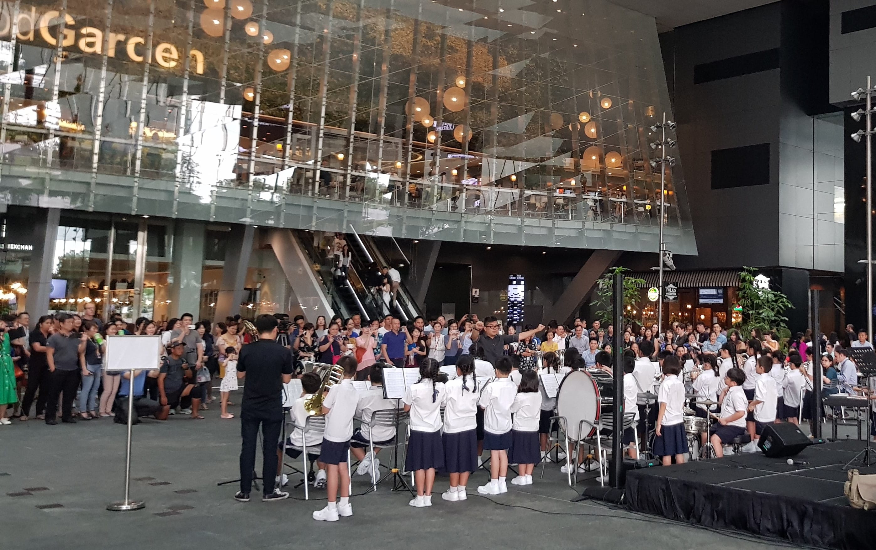 Children in white shirts perform a band concert outdoors at a mall entrance while a large crowd watches.